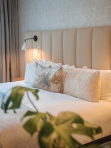 Hotel Bedroom Scene with a Beige Padded Headboard White Bedding and Decorative Embroidered Pillows a Wall mounted Lamp Lights the Bed Nearby with Curtains in the Background Oude Werf