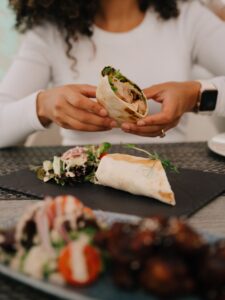 Person in a White Top Holds a Filled Tortilla Wrap Showing Greens and Chicken over a Dining Table Oude Werf