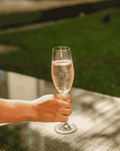 A hand holds a tall flute of sparkling wine with bubbles, outdoors on a sunlit table.
