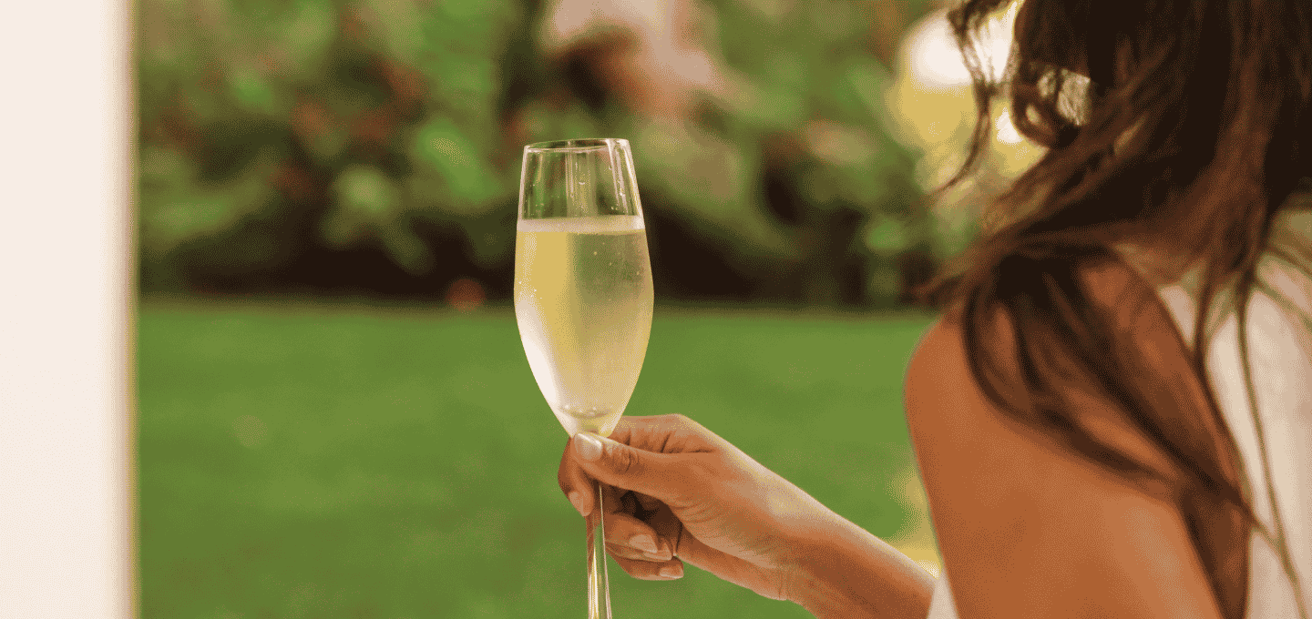 A person holding a champagne flute outdoors, with a garden scene in the background.