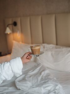 Hand in a White Robe Presenting a Glass Cup of Latte on a Saucer in a Cozy Hotel Bedroom with a Padded Headboard and Lamp in the Background Softly Lit Oude Werf